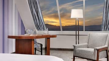 A hotel room with a bed, a table with a chair, a lamp, and a view of mountains at sunset.