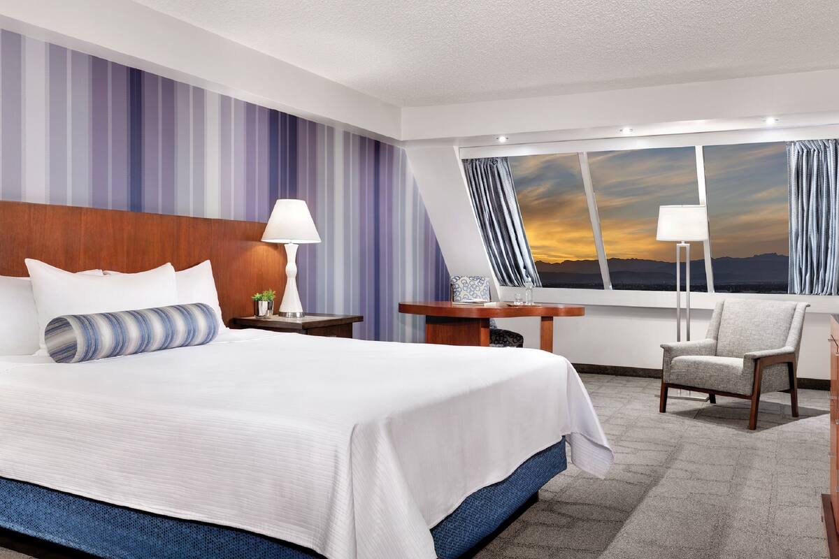 Une chambre d’hôtel avec un grand lit, un bureau, une chaise et une vue sur le coucher de soleil par la fenêtre.