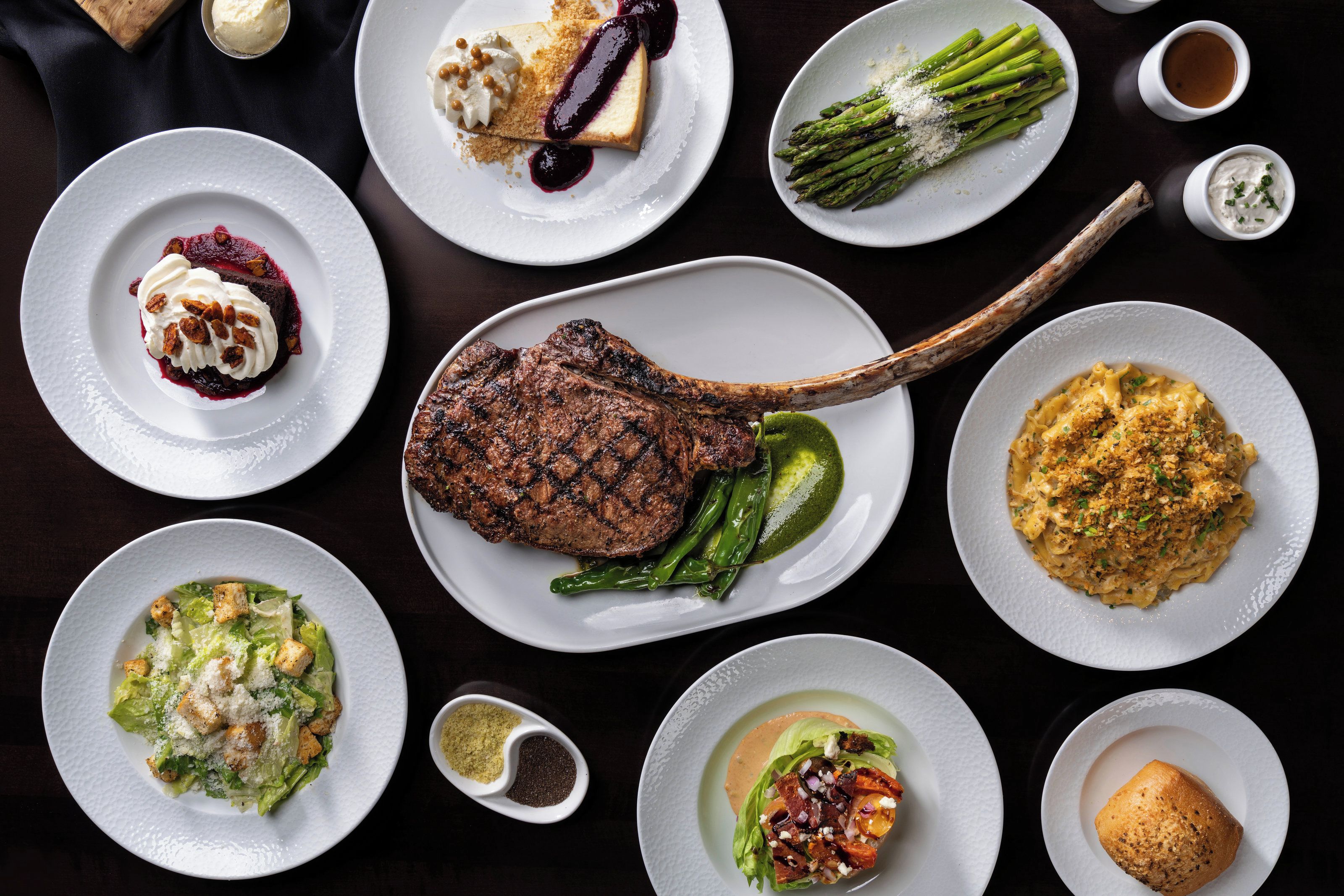 Variedad de platos que incluyen un bistec, espárragos y pasta, dispuestos sobre una mesa oscura.