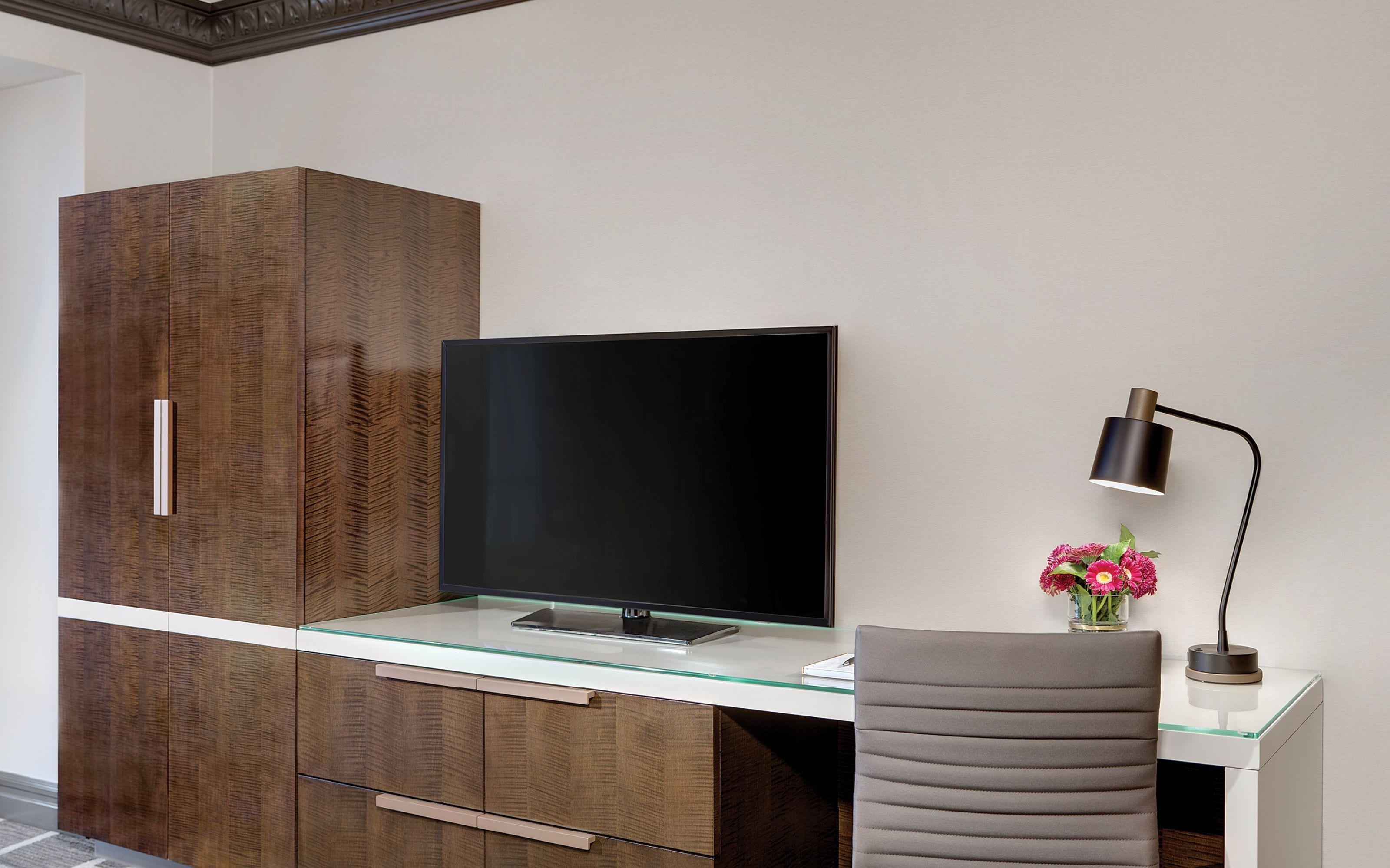 Une chambre moderne avec un téléviseur à écran plat posé sur une table en verre, une armoire en bois, une chaise grise et un bureau avec une lampe.