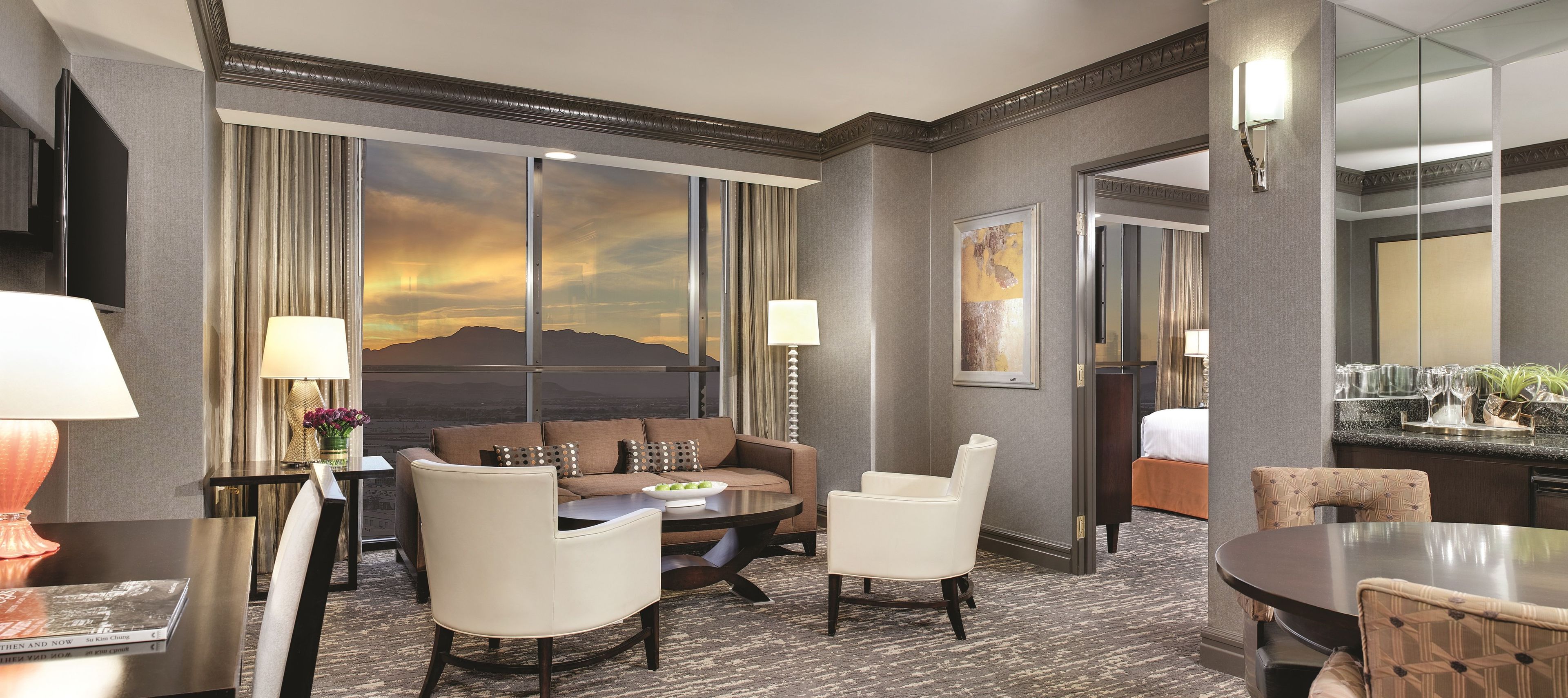 A hotel room with a large window, a sofa, two armchairs, a small table, and a dining area with a round table and chairs.