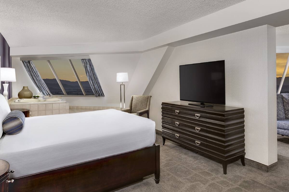 Une chambre d’hôtel comprenant un lit, une commode, une télévision, un bureau avec une lampe et une grande fenêtre donnant sur un beau paysage.