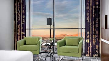 A hotel room with two green chairs, a small table, and a view of a cityscape and mountains through large windows.