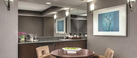 A modern hotel room with a round dining table, two upholstered chairs, a bar area with a sink, and a large mirror reflecting the room.
