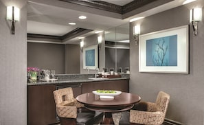 A modern hotel room with a round dining table, two upholstered chairs, a bar area with a sink, and a large mirror reflecting the room.