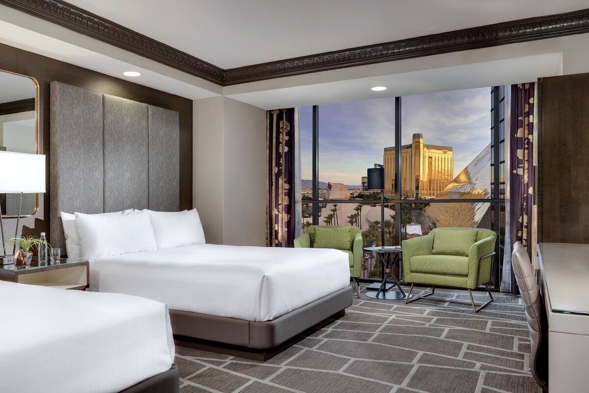 Une chambre d’hôtel avec un grand lit, deux fauteuils, un bureau et une vue sur le paysage urbain.