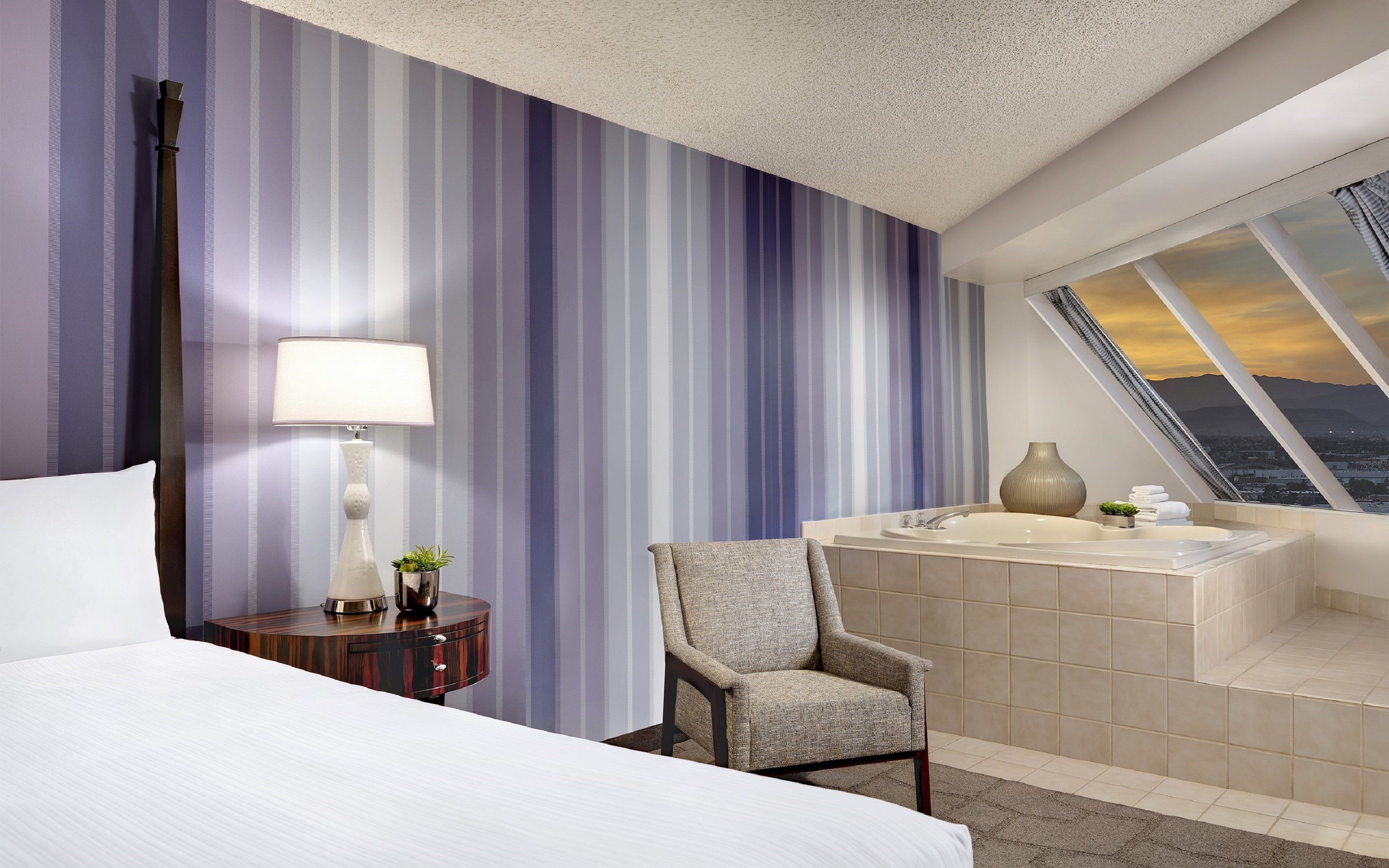 Un cuarto de hotel con cama, una silla, una mesita de noche con lámpara, un florero y vista a un atardecer a través de una ventana.