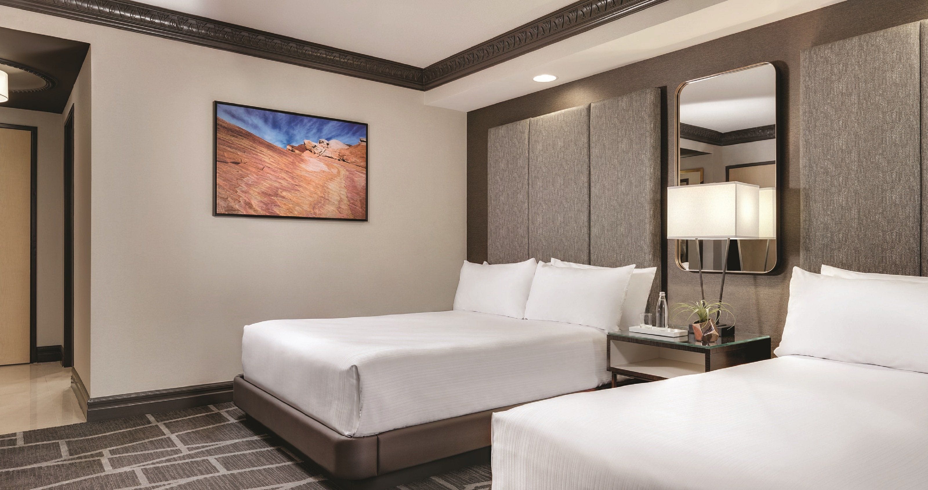 Une chambre d’hôtel avec deux lits, un grand tableau au mur, un miroir et une petite table avec une lampe.