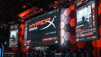 A gaming event setup with multiple screens displaying the HyperX Arena Las Vegas logo, gaming chairs, and a hexagonal wall design.