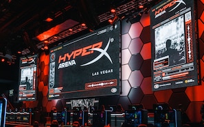 A gaming event setup with multiple screens displaying the HyperX Arena Las Vegas logo, gaming chairs, and a hexagonal wall design.