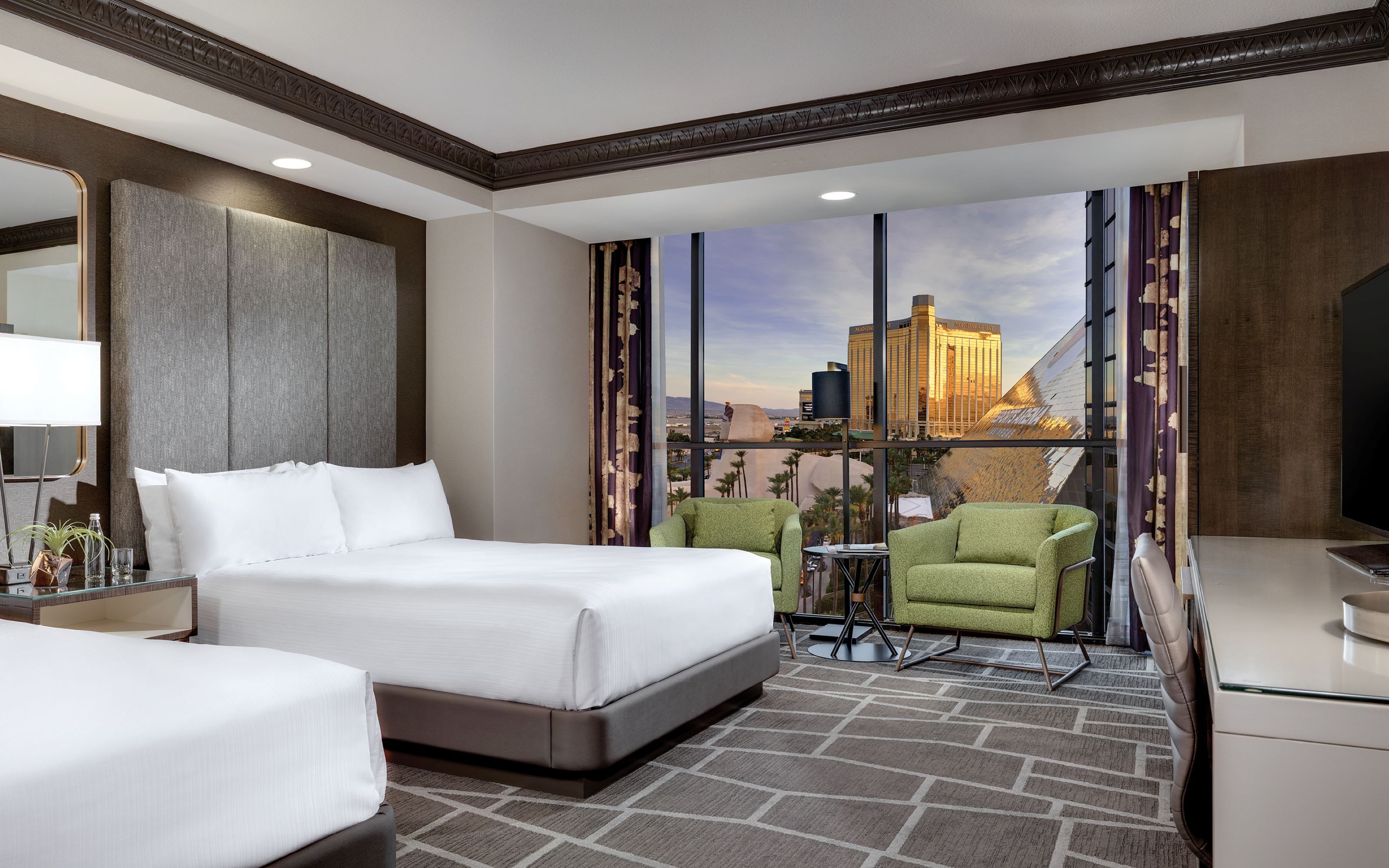 Une chambre d’hôtel avec un lit, deux chaises, un bureau et une grande fenêtre donnant sur le paysage urbain.