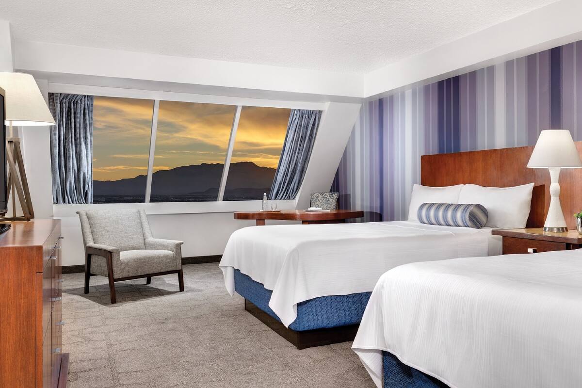 Une chambre d’hôtel avec deux lits, un bureau, une chaise, une télévision et une grande fenêtre donnant sur le coucher de soleil.