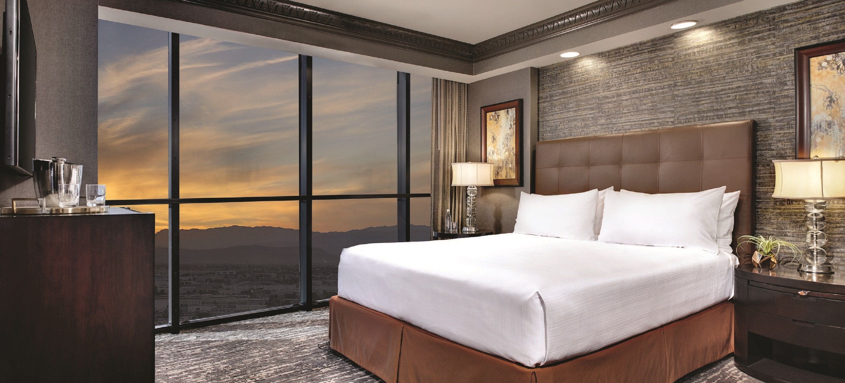 Habitación de hotel con un ventanal grande, una cama, un escritorio con lámpara y vistas a la ciudad.