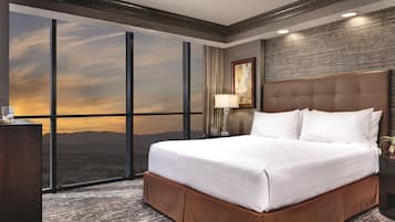 Habitación de hotel con un ventanal grande, una cama, un escritorio con lámpara y vistas a la ciudad.