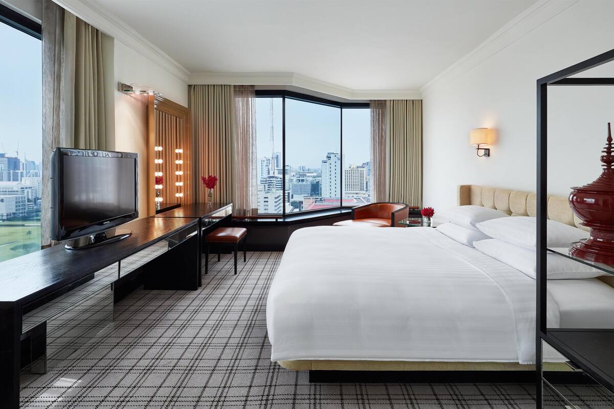 Habitación de hotel con cama, televisor, escritorio, silla, ventana grande con vista a la ciudad y una lámpara roja.