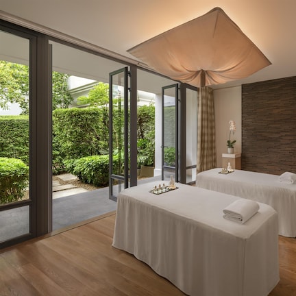 A spa room with two massage tables, a wooden wall, and a view of greenery outside.