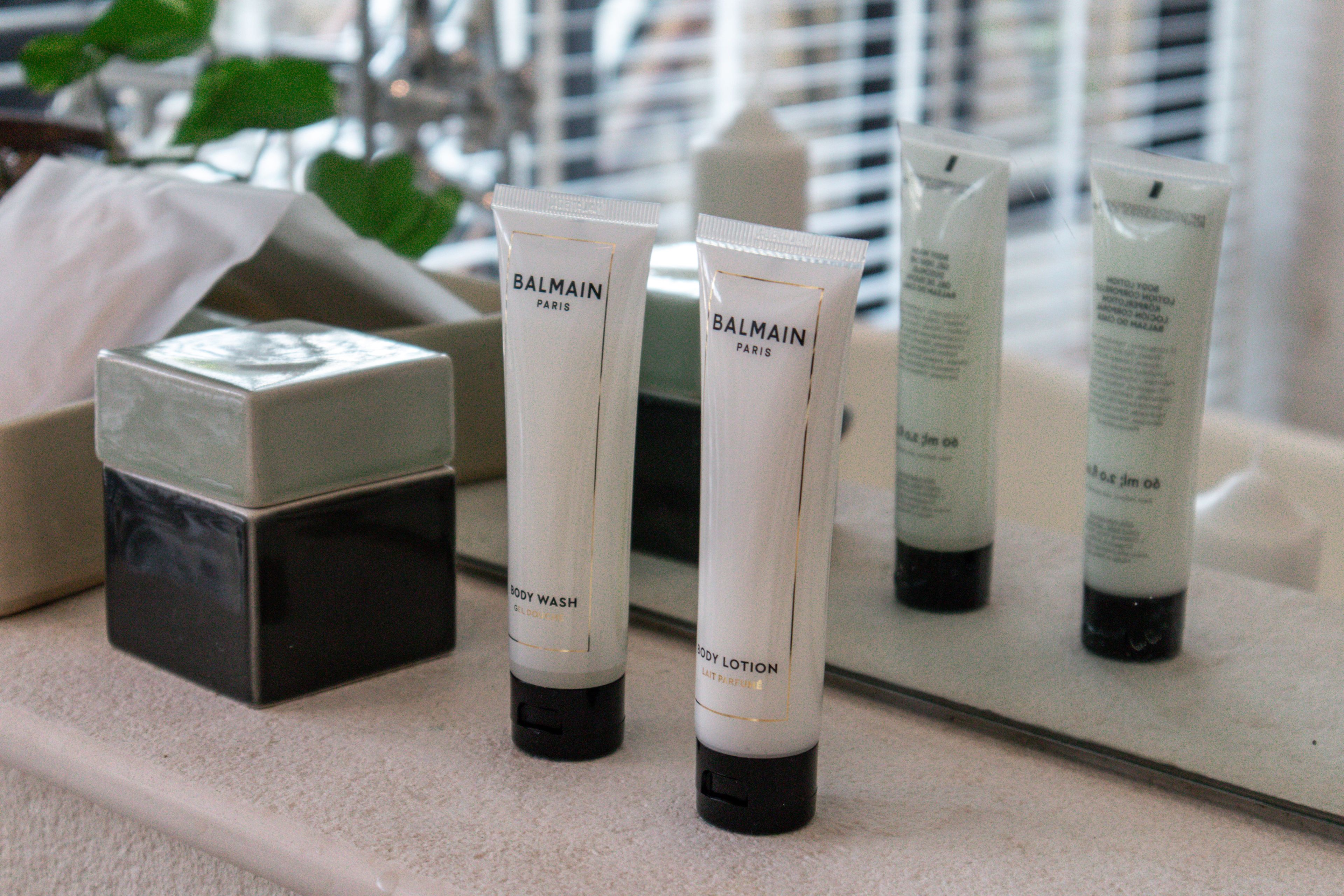 A bathroom counter with several Balmain Paris body wash and lotion tubes, a black container, and a glass jar.