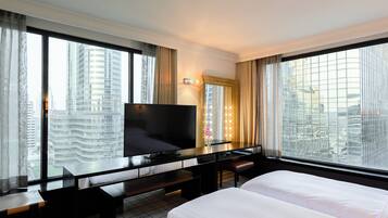 A hotel room with a large bed, a desk with a flat-screen TV, a bench, and a large window offering a city view.