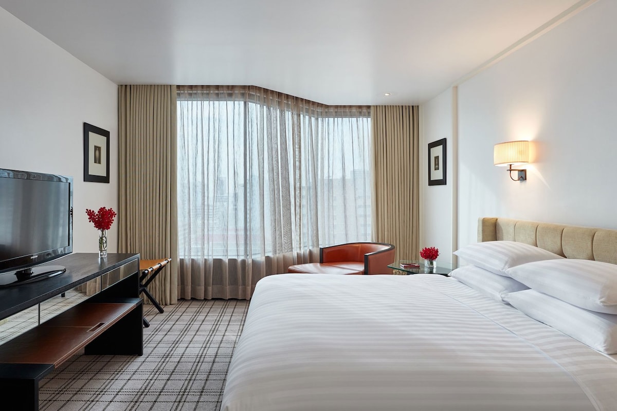Habitación de hotel con cama, escritorio con televisor, silla, lámpara y ventana con cortinas.