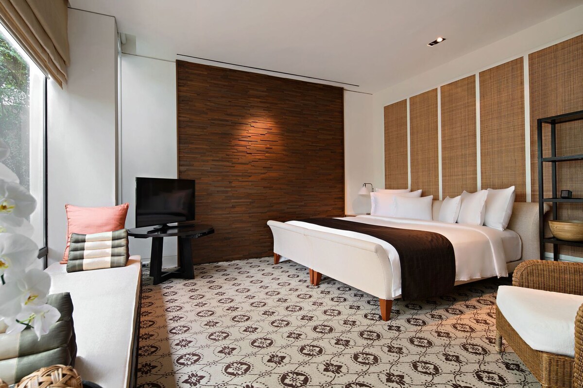 A modern hotel room with a large bed, a TV, a small table, and a seating area with a chair and a sofa.