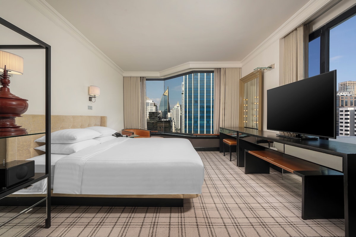 Een hotelkamer met een groot bed, een bureau met een flatscreen televisie, een stoel, een bankje en uitzicht op de skyline van de stad door een groot raam.