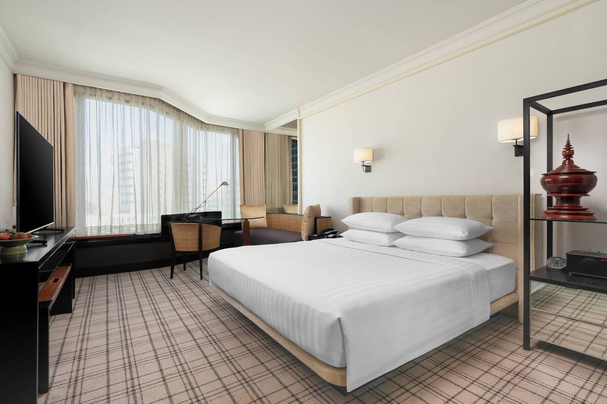 Habitación de hotel con una cama grande, un escritorio con silla, televisión y ventana con cortinas.