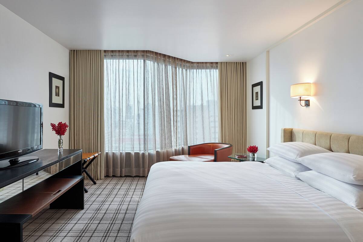 Habitación de hotel con una cama, un escritorio con televisor, una silla, una ventana con cortinas y una lámpara de pared.