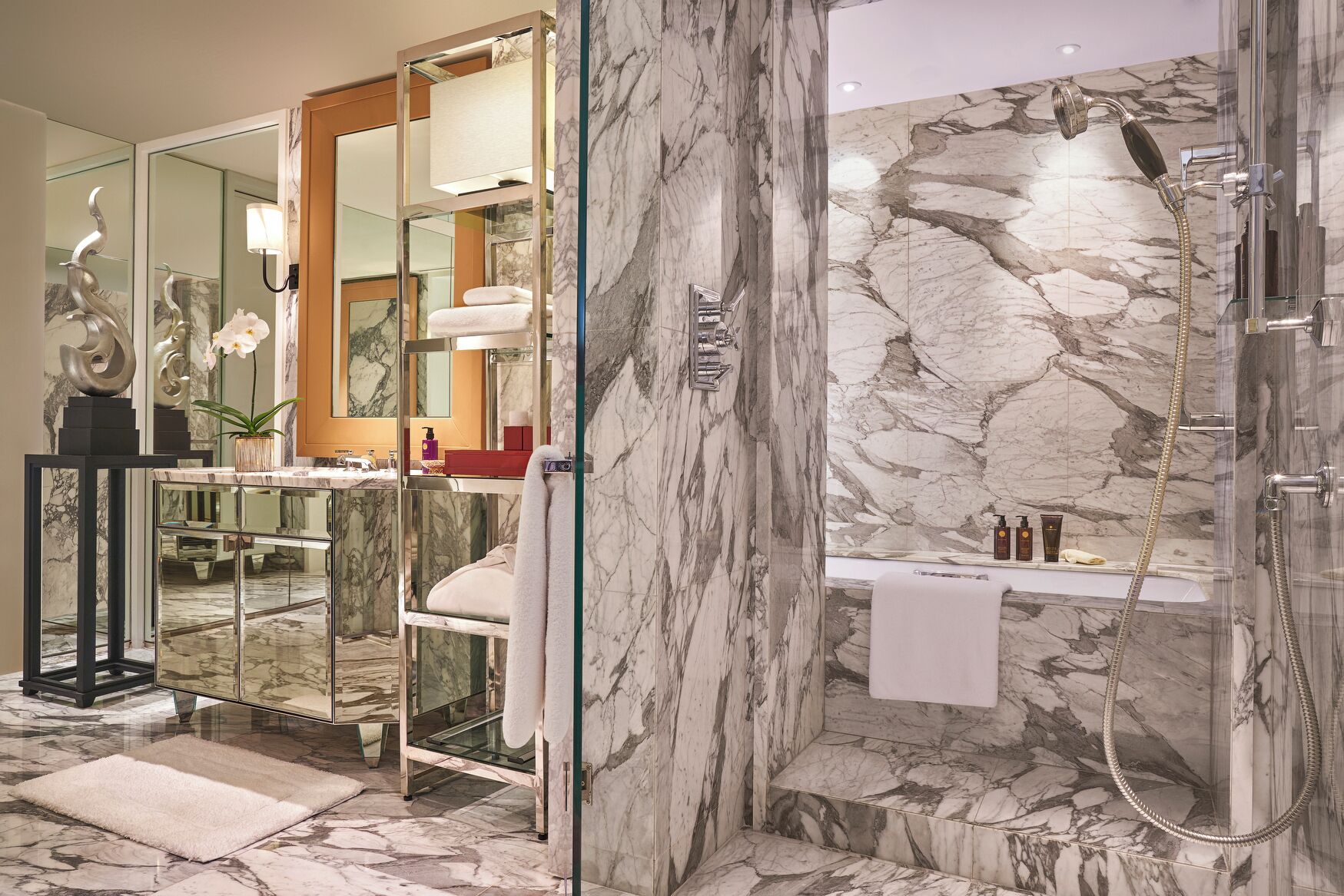 Ein luxuriöses Badezimmer mit einer Marmordusche, einem in Glas eingefassten Waschtisch und einem Spiegel mit dekorativem Rahmen.