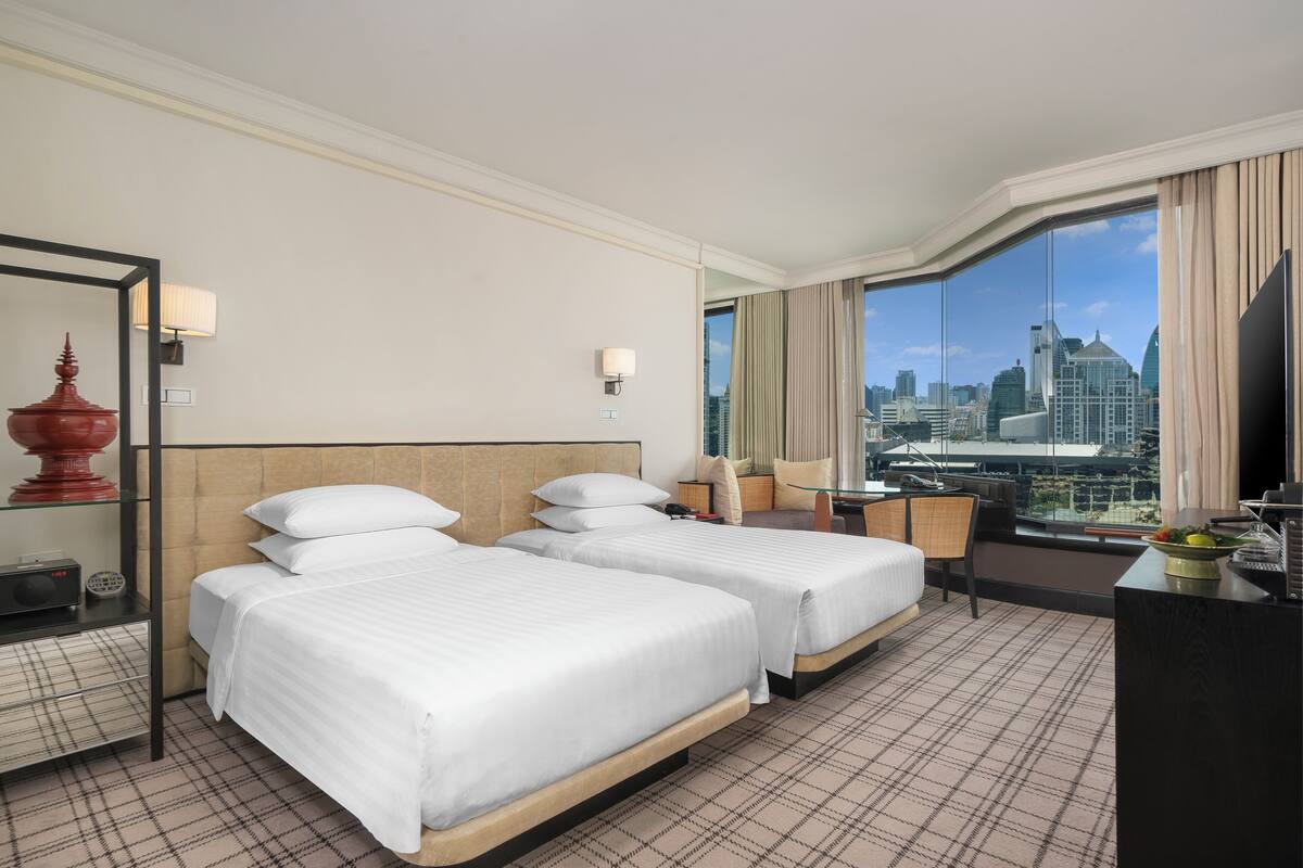 Habitación de hotel con dos camas, un escritorio, una silla, un televisor y un ventanal con vistas a la ciudad.