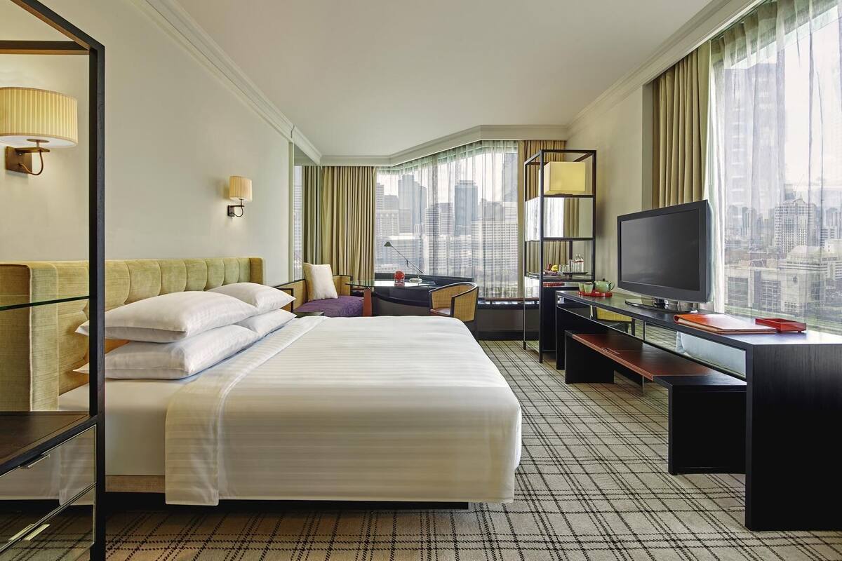 Habitación de hotel con una cama grande, un escritorio con televisor, una silla, una lámpara y vistas a la ciudad.
