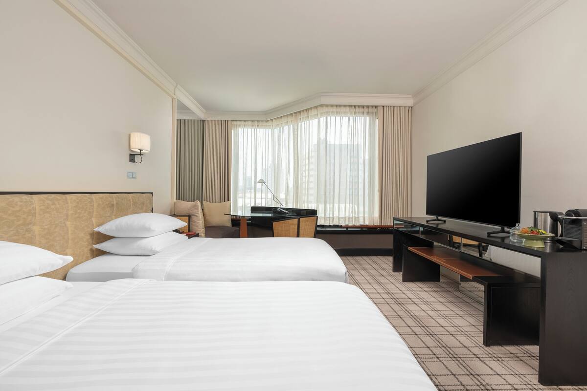 Habitación de hotel con tres camas, un escritorio, un televisor de pantalla plana, una lámpara y vistas de la ciudad a través de ventanas amplias.