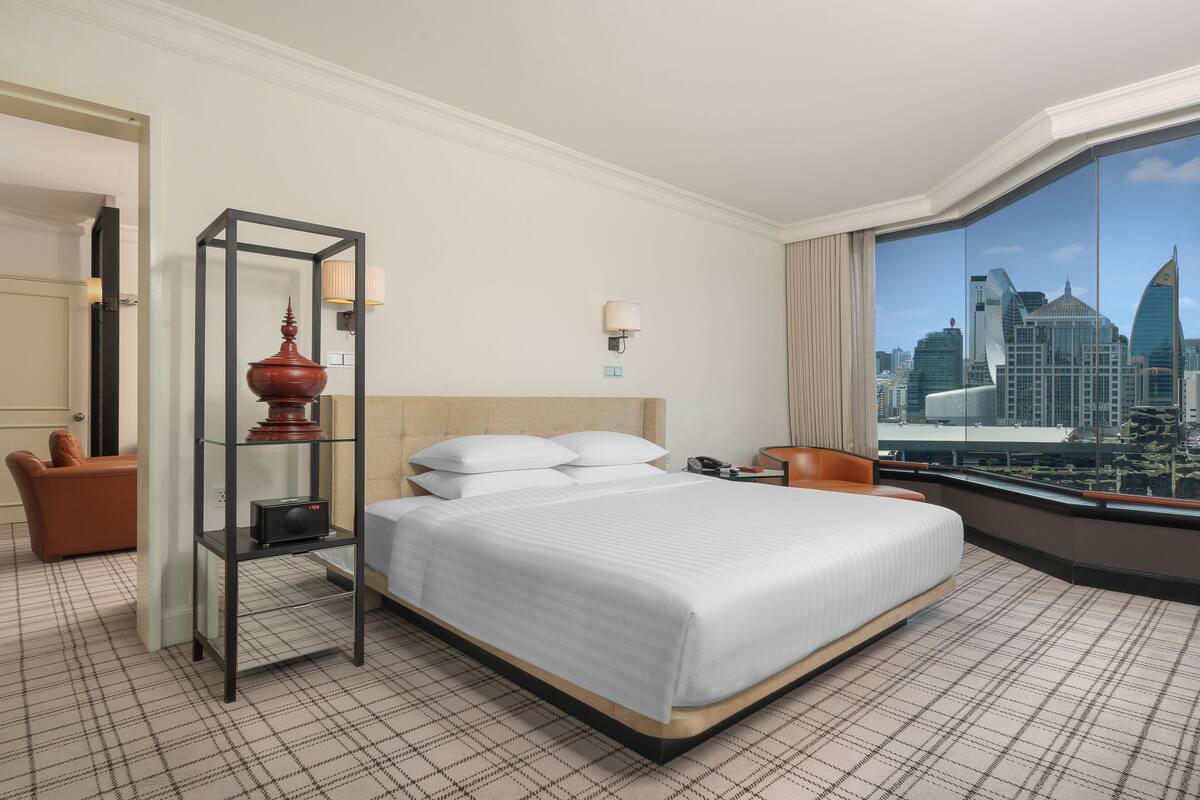 Habitación de hotel con una cama grande, un separador de vidrio, vista a la ciudad, una silla y una mesita con lámpara.
