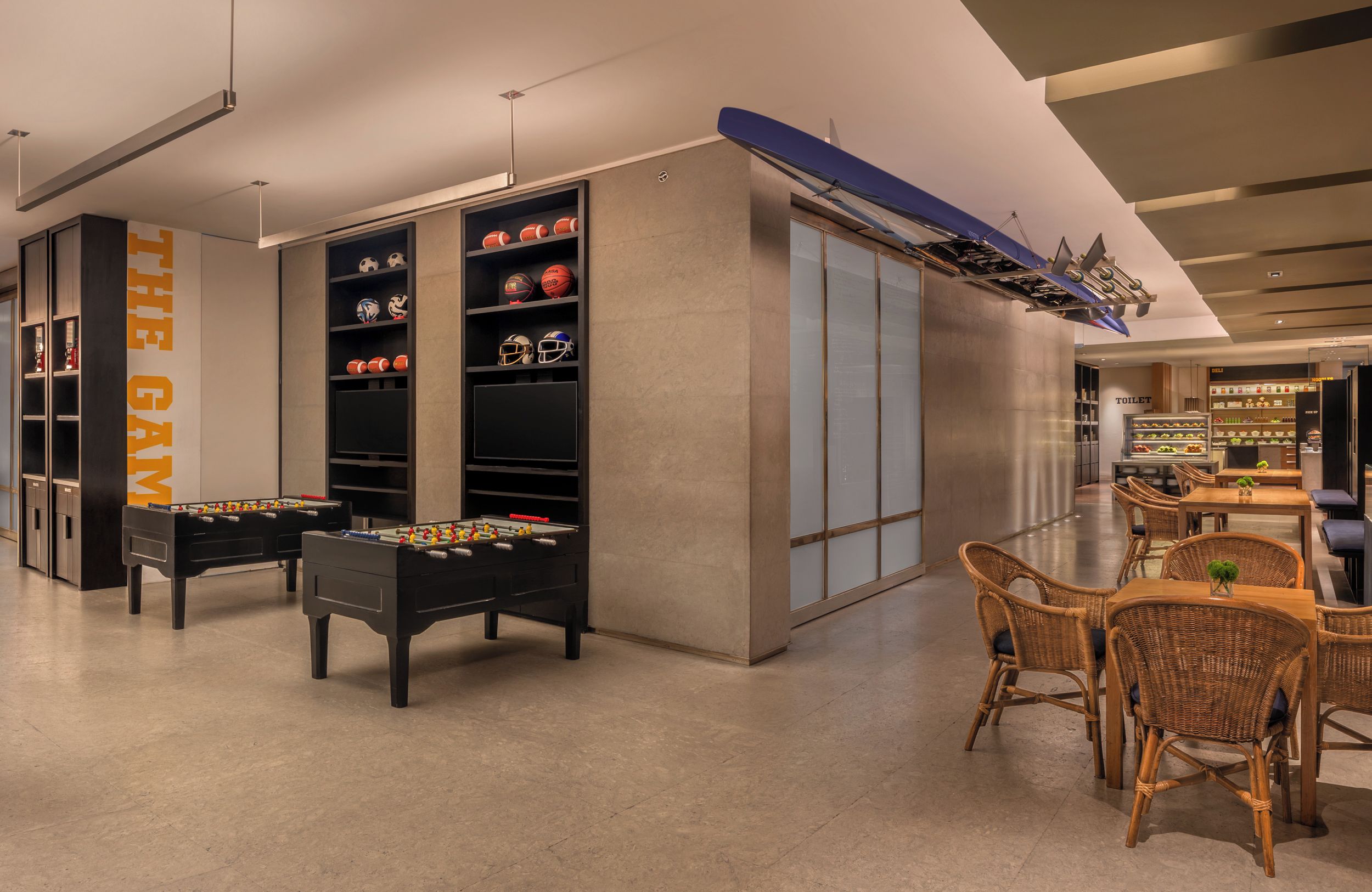 A modern indoor space with two foosball tables, a dining area with wicker chairs, and shelves with sports equipment.
