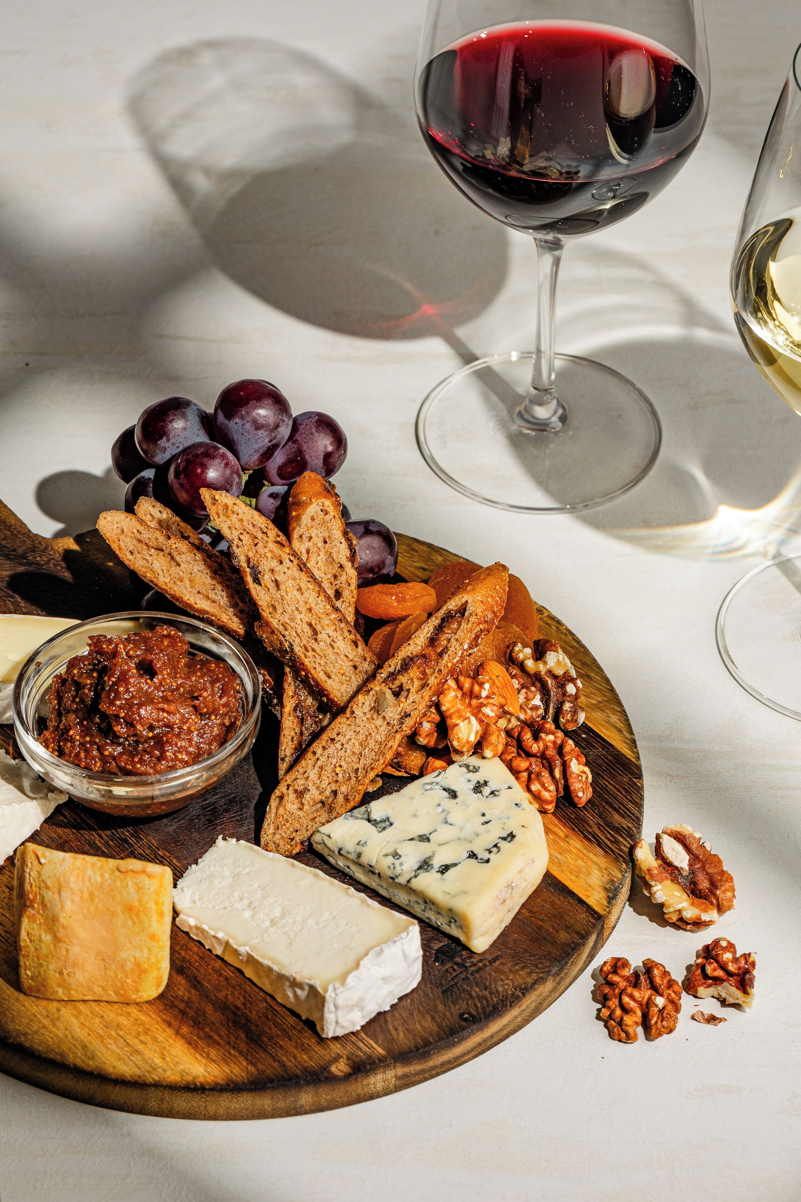 A wooden board with various cheeses, grapes, nuts, and bread, accompanied by glasses of red and white wine.