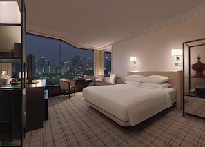 A hotel room with a large bed, a desk with a computer, a chair, a nightstand, and a view of the cityscape at night.