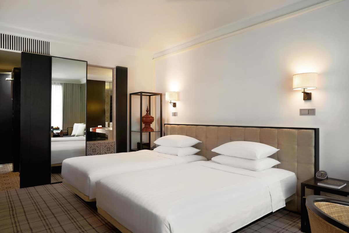 Habitación de hotel con dos camas, un espejo grande, una mesita de noche con lámpara y vistas al baño.