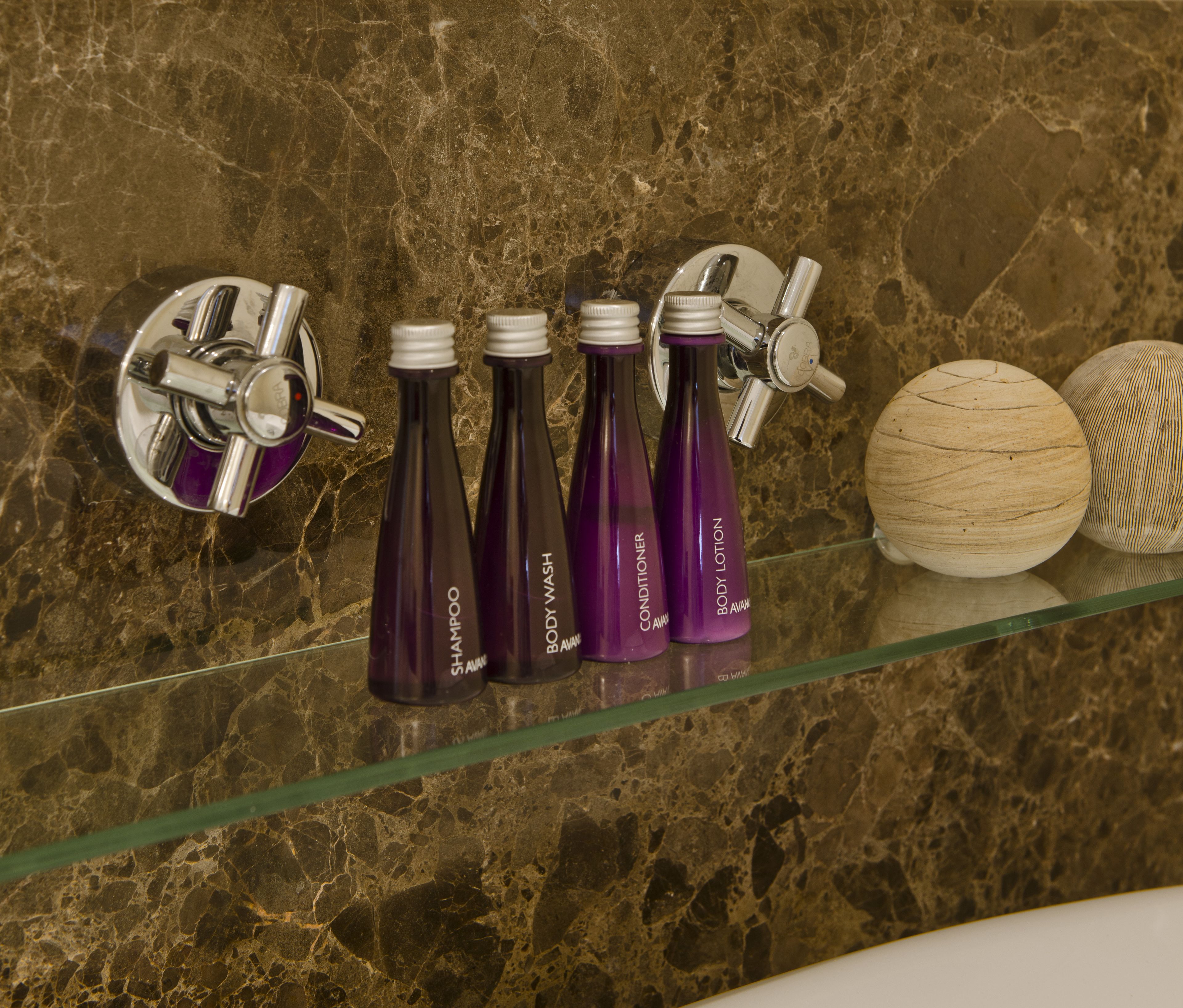 Une salle de bain avec un mur en marbre, une étagère en verre où sont posés des flacons de gel douche et d’après-shampoing, et un objet en bois.