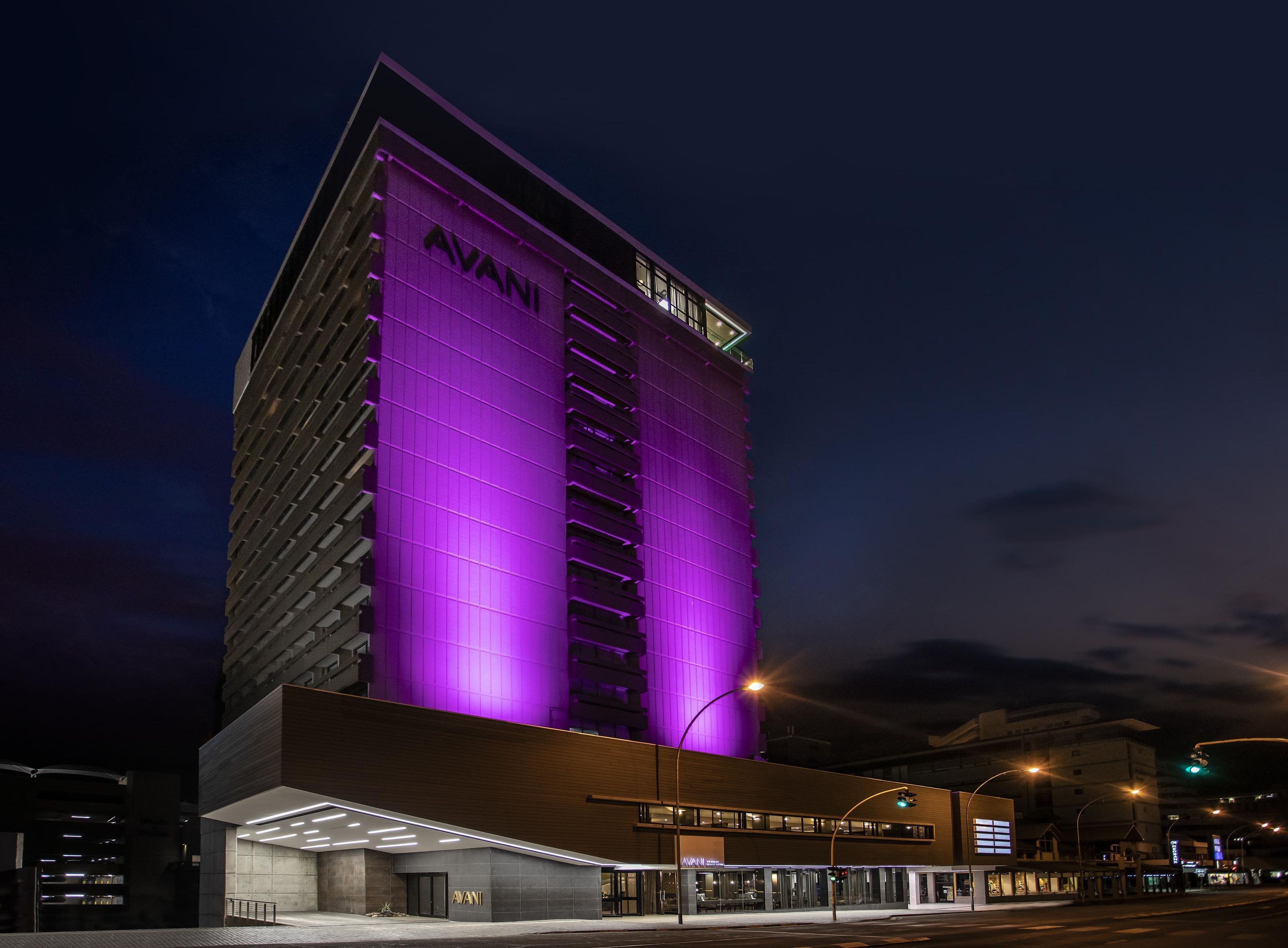 A modern building with a prominent 'AVANI' sign on the facade, featuring a unique architectural design with a purple-lit section.