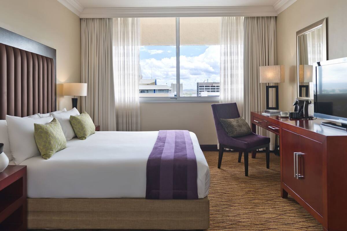 A hotel room with a bed, a desk, a chair, a TV, and a window with a city view.