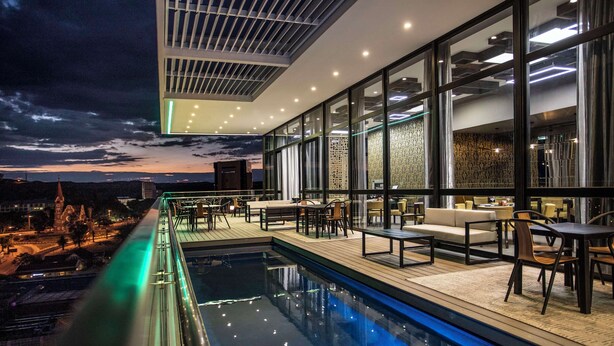 Zona de piscina en la azotea con mobiliario exterior y vistas a la ciudad al anochecer.