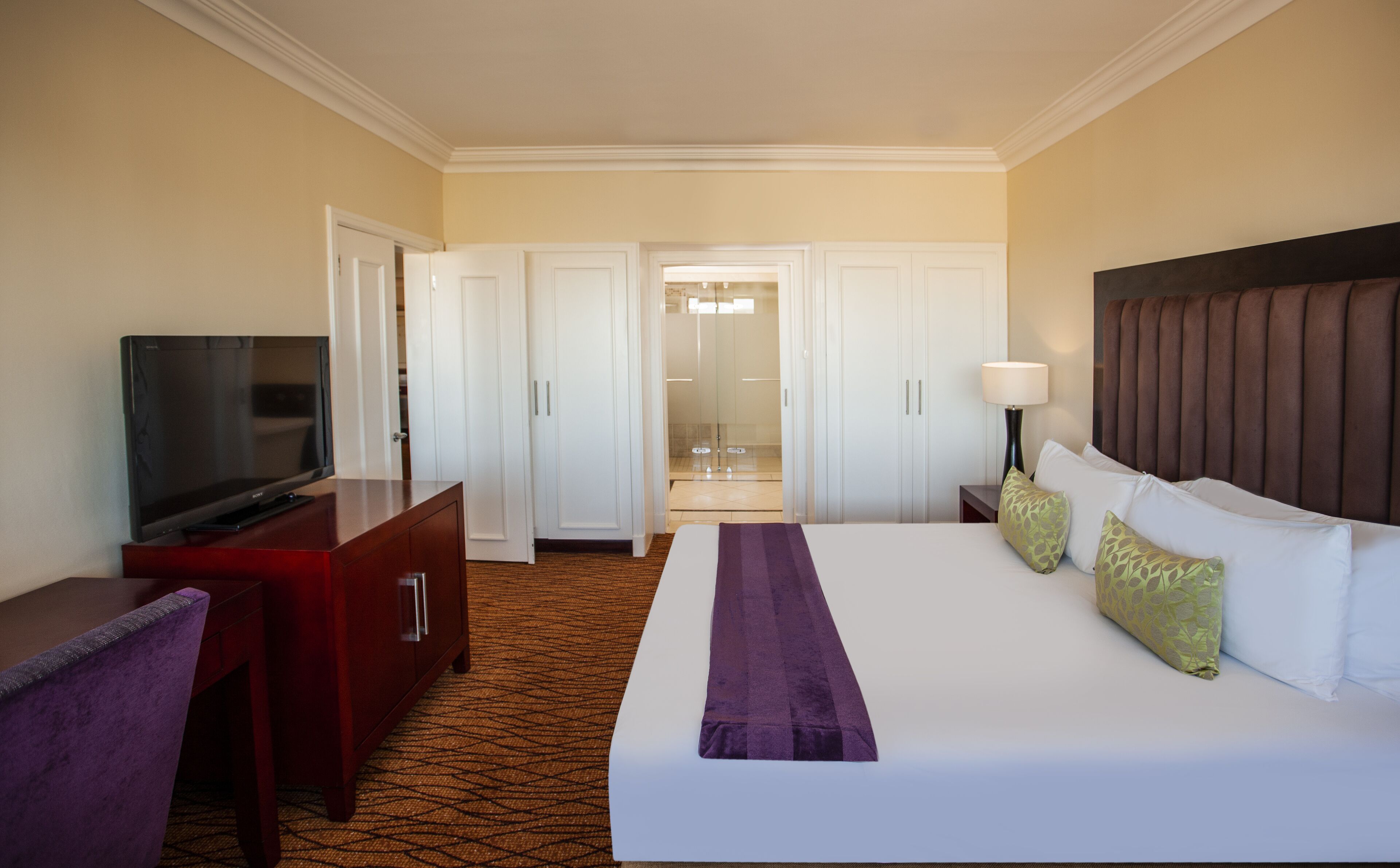 Habitación de hotel con cama, televisor, escritorio y baño visible a través de una puerta abierta.
