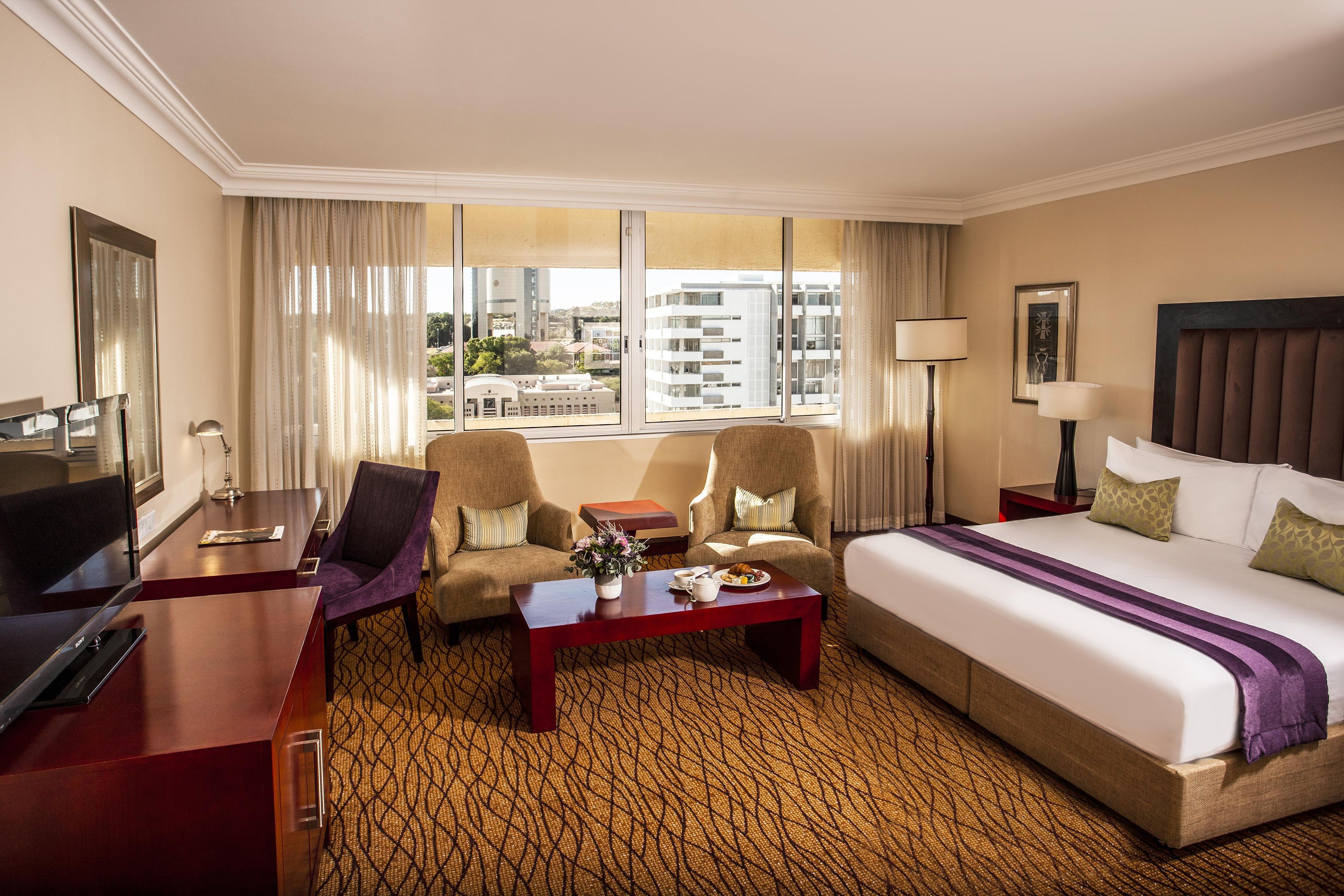 A hotel room with a bed, two armchairs, a desk, a coffee table, and a view of buildings through large windows.