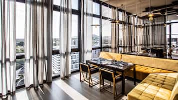 Un comedor moderno con sofá, mesa y sillas, ventanas amplias con cortinas e iluminación colgante.