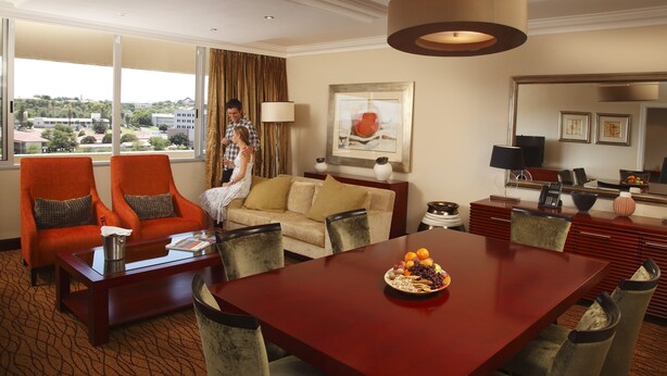 Una habitación de hotel con comedor, un sofá, dos sillones, un ventanal con vistas a edificios y un cuadro en la pared.