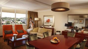 A hotel room with a dining area, a sofa, two armchairs, a large window with a view of buildings, and a painting on the wall.