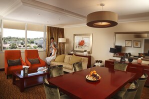 Quarto de hotel com área de refeições, sofá, duas poltronas, uma janela grande com vista para edifícios e um quadro na parede.