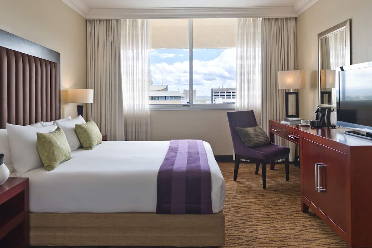 A hotel room with a large bed, a desk with a TV, a chair, and a view of buildings through the window.