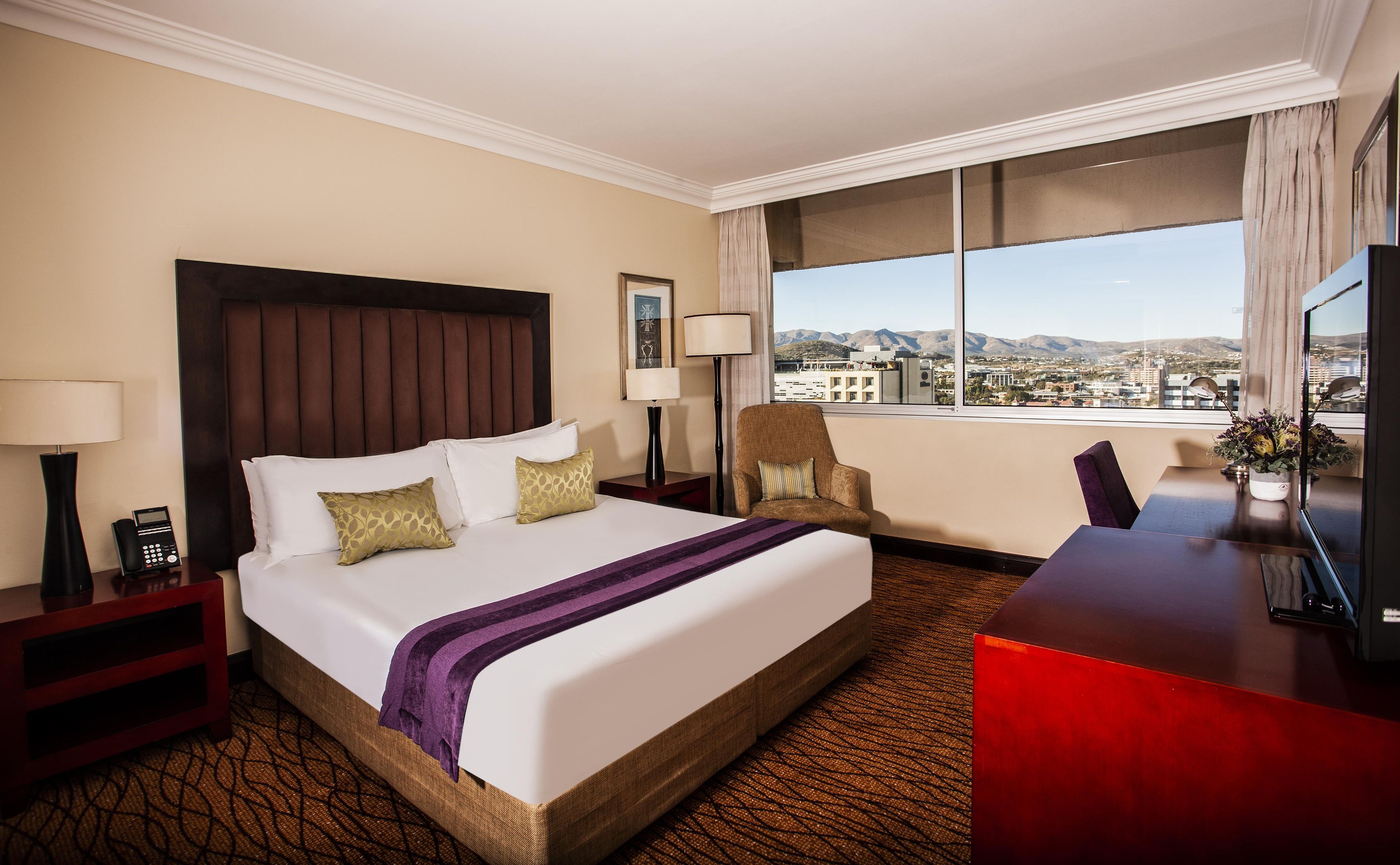 Habitación de hotel con una cama grande, un escritorio, una silla, una lámpara, un teléfono y vistas a la ciudad.