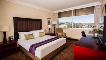 A hotel room with a large bed, a desk, a chair, a lamp, a phone, and a view of the city.