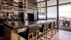 Una zona de bar moderna con una barra alargada, varios taburetes y vistas a la ciudad a través de grandes ventanales.